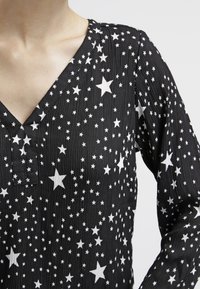 Schwarze Bluse mit V-Ausschnitt und langen Ärmeln, die mit weißen Sternmustern verziert ist. Der Stoff hat eine strukturierte, zerknitterte Optik.