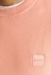 Pull rose pâle texturé avec un col rond et un petit patch carré portant l'inscription "BOSS" dans le coin inférieur.