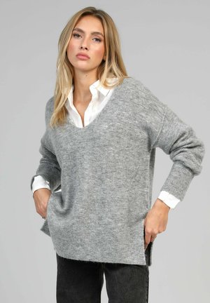 Jeune femme portant un pull ample gris par-dessus une chemise blanche, debout les mains dans les poches devant un fond gris clair uni.