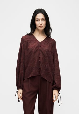 Femme portant un chemisier texturé marron rouge à boutons avec des manches longues et un pantalon assorti de la même couleur marron rouge, devant un fond blanc uni.