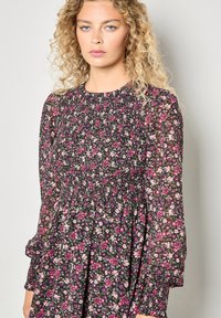 Czarna sukienka w kwiaty z marszczonym stanikiem, długimi przezroczystymi rękawami oraz mieszanką różowych i fioletowych kwiatów na ciemnym tle.