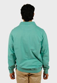 Pullover turchese con maniche lunghe, polsini e orlo a coste. Texture morbida e scollo colletto. Visto da dietro, indossato con pantaloni beige.