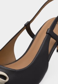 Κοντινή λήψη μαύρου δερμάτινου παπουτσιού slingback με ταμπά εσωτερική σόλα και διακοσμητική αγκράφα ανοιχτού χρώματος στο μπροστινό λουράκι.