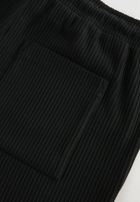 Pantalones jogger de tela negra acanalada con un gran bolsillo lateral, cintura elástica, rayas verticales y textura suave.