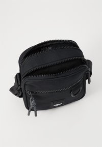 Sort stof crossbody tas med to lynlåslommer, tekstureret overflade og vævet hardware-accent. Har justerbar strop og logo-mærke.