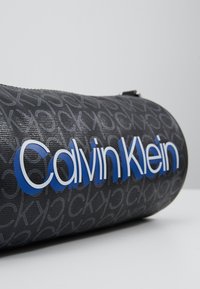Borsa cilindrica nera con motivo a rilievo del logo grigio e scritta blu "Calvin Klein". Materiale testurizzato con finitura liscia.