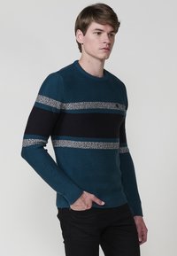 Suéter de punto en verde azulado con detalles de rayas en azul marino y gris. Diseño de cuello redondo, puños y dobladillo acanalados, y una tela texturizada.