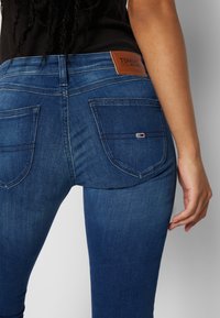 Jeans de mezclilla azul medio con un diseño ajustado, que incluyen bolsillos traseros y un parche de cuero marrón etiquetado como "TOMMY JEANS". Textura suave.
