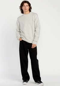 Pull tricot gris clair avec un col rond et des poignets côtelés, associé à un pantalon large noir et des baskets noires. Fond neutre.