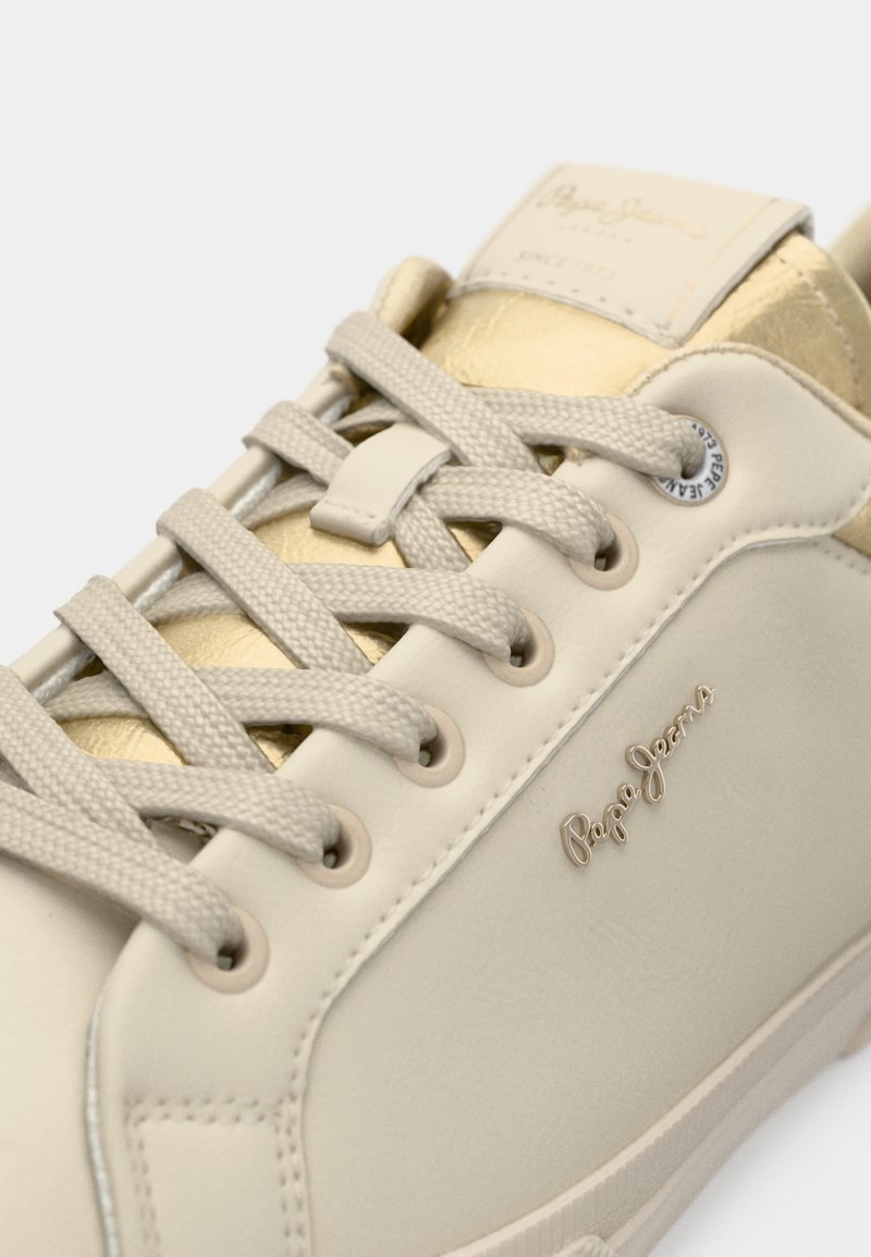 Zapatillas de cuero beige con detalles dorados, cordones planos y logo de la marca. Textura suave y diseño elegante con detalles de color contrastantes.