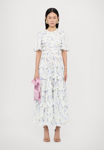 Witte maxi-jurk met bloemenmotief en gegolfde lagen, met blauw en gele bloemen. Gecombineerd met een lichtroze handtas en beige sandalen.