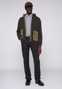 Cárdigan de punto gris oscuro con detalles en verde oliva y dos bolsillos frontales, superpuesto a una camisa a rayas azul y blanca y pantalones grises.