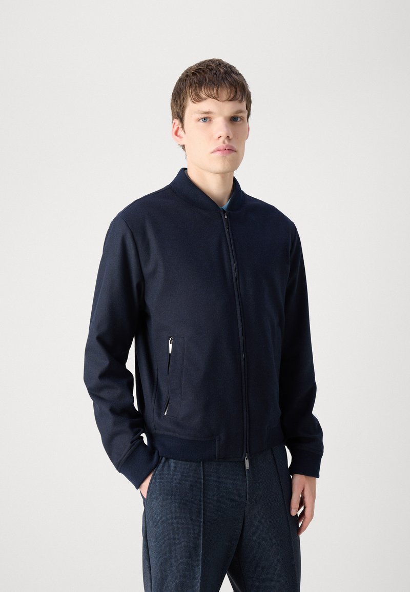 Granatowa kurtka bomberka zapinana na zamek z kołnierzem stójkowym, ściągaczami przy mankietach i dwoma bocznymi kieszeniami. Wykonana z teksturowanej tkaniny, ma luźny krój.