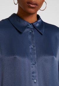 Marineblauw satijnen shirt met een puntkraag, sluiting met knopen aan de voorkant en een gladde textuur. Voorzien van bijpassende knopen en een klassiek ontwerp.