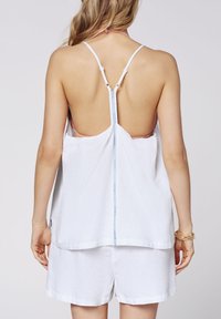 White sleeveless top with a low back and adjustable straps, featuring a blue accent piece at the center. Combined with white shorts.