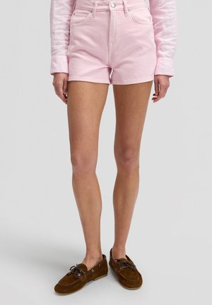 Mujer con pantalones cortos de talle alto rosa, camiseta de manga larga rosa claro y mocasines de ante marrones, de pie sobre un fondo claro.