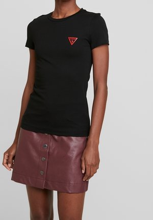 Kobieta ubrana w dopasowany czarny t-shirt z krótkim rękawem, z czerwonym logo w kształcie trójkąta, oraz mini spódniczkę z bordowej skóry zapinaną na guziki z przodu.