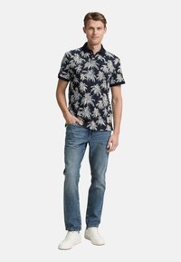 Polo de manga corta en azul marino con un patrón de palmeras de color claro. Combinado con jeans azul claro y zapatillas blancas.