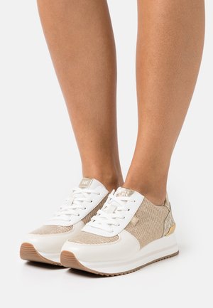 Baskets en maille blanc et beige avec des accents dorés, portées sur des jambes nues, vues de face et de profil sur un fond blanc.