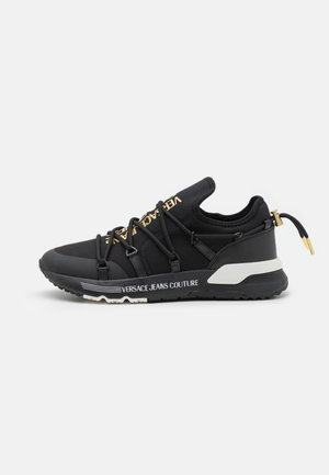 Sort Versace Jeans Couture sneaker med guldlogo på tungen, sorte snøreløkker, hvide detaljer på sålen og trækflap med gyldne ender ved hælen.