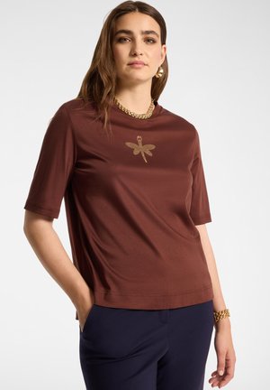 Mujer vestida con una camiseta marrón de manga corta con diseño de libélula dorada, pantalones azul marino, collar de cadena dorada, pulsera y pendientes, de pie con la mano en el bolsillo.