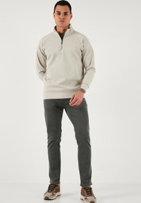 Svjetlosmeđi dukserica s četvrtim zatvaračem i raglan rukavima, u kombinaciji s tamnosivim uskim hlačama i višebojnim tenisicama. Jednostavni dizajn, bez vidljivih uzoraka.