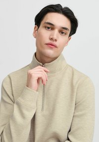 Beige zip-ups genser med høy krage og lange ermer, med en minimal tekstur og en liten glidelås detalj ved halsen.