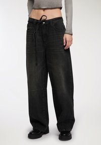 Osoba w szerokich, wyblakłych czarnych dżinsach z wiązanym paskiem w talii, skróconej szarej bluzie z długim rękawem i czarnych butach na grubej podeszwie.