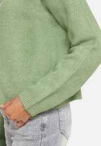 Zielony sweter z dzianiny o fakturowanej powierzchni i ściągaczami przy mankietach, zestawiony z jasnoszarymi dżinsami. Sweter ma prosty, pudełkowaty krój i luźne dopasowanie.