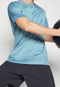 Hellblaues, feuchtigkeitstransportierendes Sport-T-Shirt mit kurzen Ärmeln, das ein dezentes Logo auf der Brust trägt, kombiniert mit dunklen Shorts.