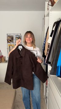 Žena s blond vlasy v bílé košili a džínách, která drží tmavě hnědou sako na knoflíky, uvnitř místnosti s oblečením a časopisy.