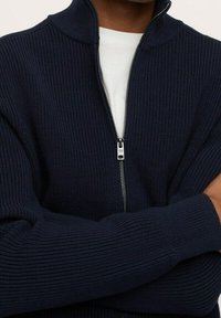Mann in einem navyblauen, gerippten Reißverschluss-Pullover über einem weißen Hemd, mit verschränkten Armen, der Reißverschluss auf Brusthöhe teilweise geöffnet.