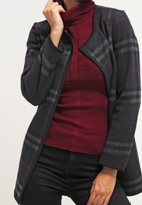 Giacca a scacchi nera e grigia con maniche lunghe, tessuto texturizzato, apertura frontale, indossata sopra un maglione a collo alto ribbed bordeaux.