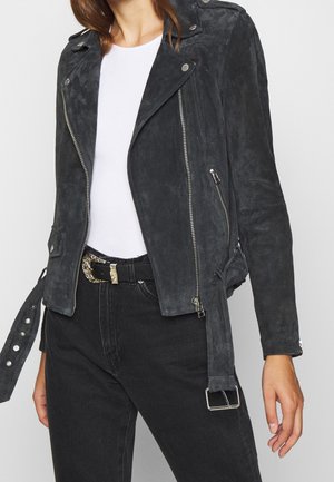 Nainen, jolla on musta mokkanahkainen motoristintakki hopeisilla vetoketjuilla, valkoinen paita, mustat korkeavyötäröiset farkut ja musta vyö koristeellisella soljella.