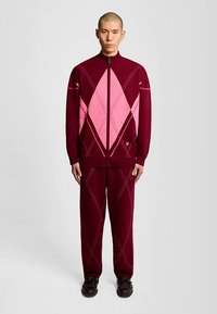 Camisola de malha com fecho de correr em borgonha com padrão de losangos cor-de-rosa, punhos com costura canelada, e calças a combinar. Ambos feitos de um tecido texturado. Sapatos pretos usados.