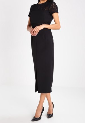 Femme portant une robe midi noire ajustée avec des manches courtes en dentelle et une fente avant, accompagnée de talons hauts noirs à bout pointu.