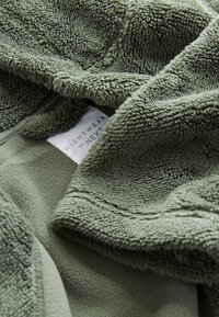 Tissu polaire vert avec une surface douce et texturée, comprenant une étiquette indiquant « Nightwear by Next » intégrée dans les plis du matériau.