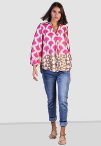 Farbige, lockere Bluse mit pinkem Blumenmuster und braunen geometrischen Akzenten, kombiniert mit hellblauen, ausgewaschenen Jeans und braunen Sandalen.