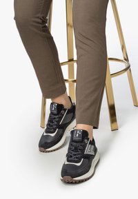 Zapatillas deportivas en negro y gris con una parte superior texturizada, acentos reflectantes y suelas blancas gruesas, combinadas con pantalones marrones ajustados.