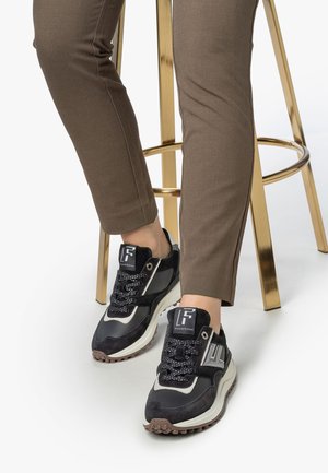 Zwarte en grijze sportieve schoenen met een gestructureerde bovenkant, reflecterende accenten en dikke witte zolen, gecombineerd met aansluitende bruine broeken.