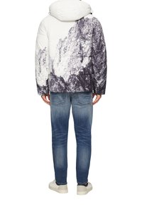 Pufferjacke mit weißem und schwarzem Bergdruck, Reißverschluss, Kapuze und gestepptem Textil, kombiniert mit hellblauen Jeans und weißen Turnschuhen.