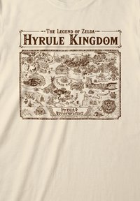 Krämfärgad t-shirt med en tryckt karta över Hyrule kungarike i brunt. Designen inkluderar berg, skogar och landmärken med detaljerade illustrationer.