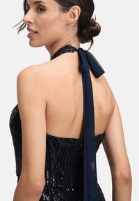 Kobieta ubrana w ciemnoniebieską, cekinową sukienkę na ramiączkach z kokardą z tyłu, z włosami ułożonymi w niskiego koka, patrząca na bok.