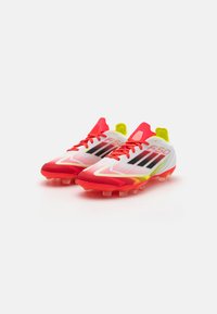 White and red football boots with a mesh upper, featuring black stripes, neon yellow accents, and a textured sole with conical studs.