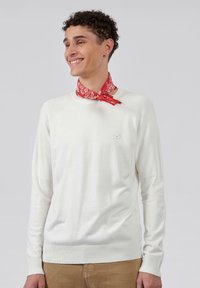 Pull en maille blanche avec des accents côtelés sur les épaules et les manches, arborant un petit logo sur la poitrine. Associé à un bandana rouge à motifs.