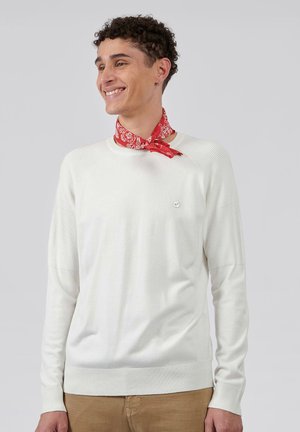 Witte gebreide trui met geribbelde accenten op de schouders en mouwen, met een klein logo op de borst. Gecombineerd met een patroon van rode bandana.