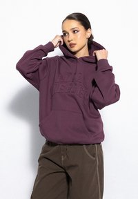Fioletowa bluza z kapturem z wytłoczonym napisem "LOCAL HEROES", kieszenią z przodu i luźnym fasonem, zestawiona z ciemnobrązowymi spodniami.