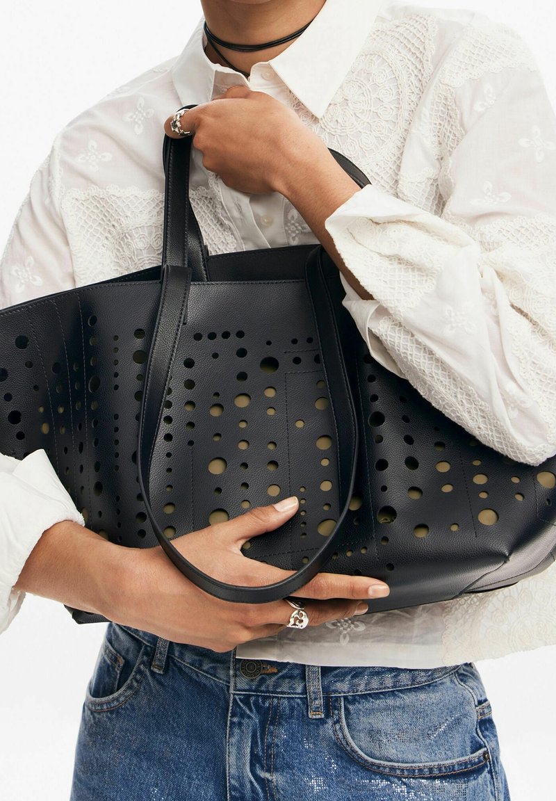 Sac fourre-tout noir perforé avec doubles poignées, présentant des découpes circulaires et une texture lisse, tenu par une personne portant une chemise blanche brodée.