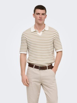 Młody mężczyzna w beżowo-białej prążkowanej dzianinowej koszulce polo z krótkim rękawem, beżowych spodniach i ciemnobrązowym pasku, stojący z jedną ręką w kieszeni.