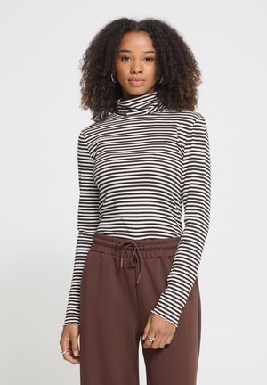 Femme debout, de face, portant un col roulé rayé noir et blanc et un pantalon marron taille haute à cordon, sur fond uni.